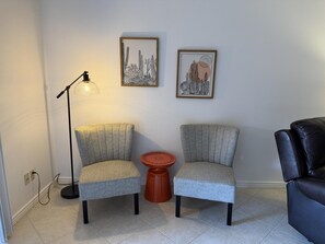 A room with two grey armchairs, a floor lamp, a small orange table, and two framed artworks on the wall.