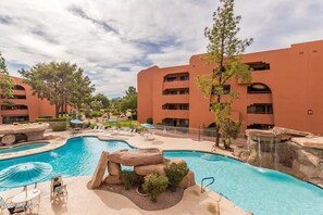 Un hotel con una amplia piscina exterior, varios balcones y una zona ajardinada con árboles y rocas.