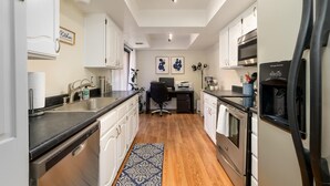 Una cucina moderna con mobili bianchi, un piano di lavoro nero, lavastoviglie integrata, frigorifero e forno a microonde. Il pavimento è in legno e c'è un tappeto azzurro con decorazioni.