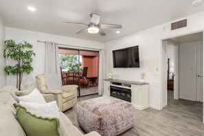 A living room with a sofa, ottoman, ceiling fan, flat-screen TV, and a fireplace. - Casa Amor - Anasazi Village - Full Golf Course Views (Phoenix, Arizona)