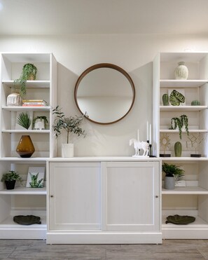 A white interior with a round mirror above a white cabinet, surrounded by shelves with various plants and decorative items.