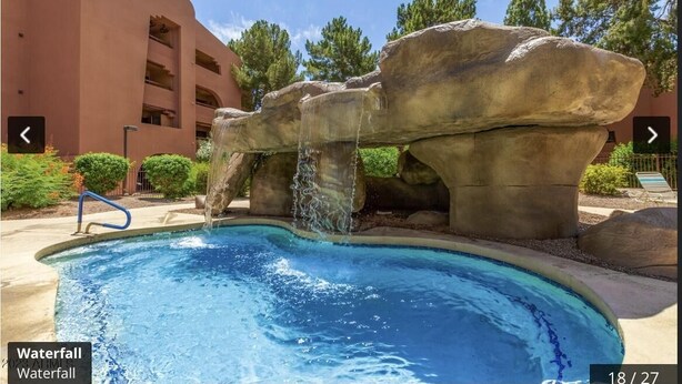 Una piscina con caída de agua, rodeada de grandes formaciones rocosas y un edificio con balcones.