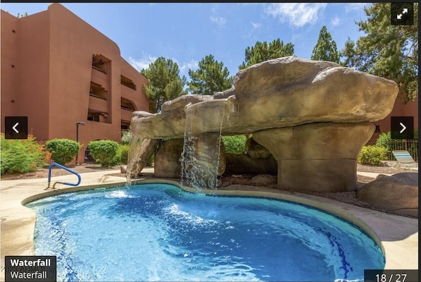A pool with a waterfall feature, surrounded by large rock formations and a building with balconies.
