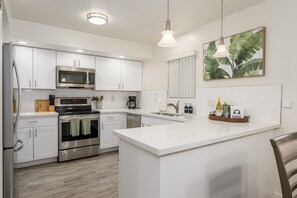 A modern kitchen with white cabinetry, stainless steel appliances, a central island, and a large window with blinds. - Casa Amor - Anasazi Village - Full Golf Course Views (Phoenix, Arizona)