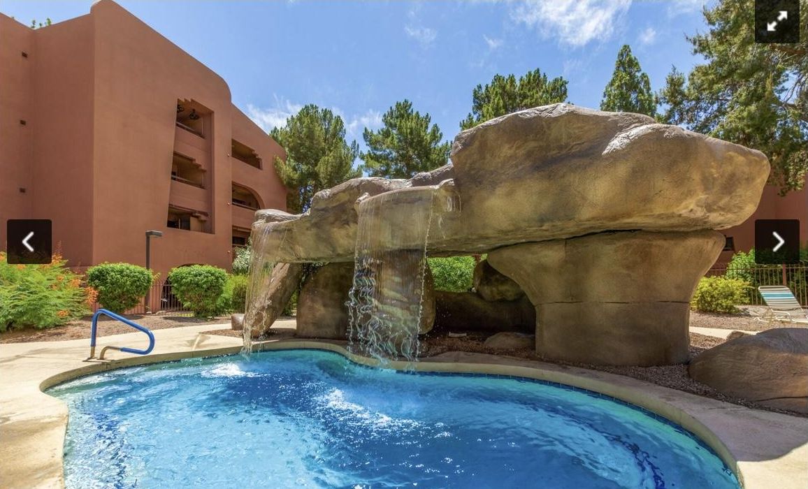 A pool with a waterfall feature, surrounded by large rock formations and a building with balconies.