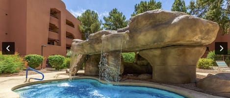 A pool with a waterfall feature, surrounded by large rock formations and a building with balconies.