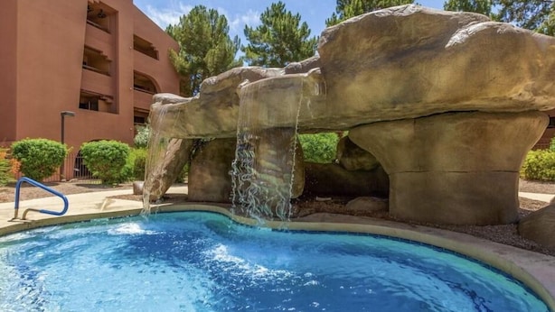 A pool with a waterfall feature, surrounded by large rock formations and a building in the background. - CASA ANA - Anasazi Village - Updated unit, pool view. (Phoenix)