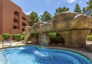 A pool with a waterfall feature, surrounded by large rock formations and a building in the background.
