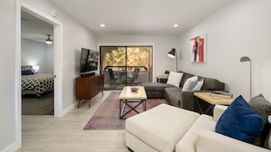 A modern living room with a sofa, a coffee table, and a TV. There is a balcony with a view of trees. - CASA ANA - Anasazi Village - Updated unit, pool view. (Phoenix)