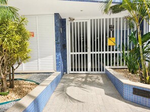 Uma entrada protegida por portão, com um caminho de azulejos azuis, uma palmeira e uma placa indicando área de estacionamento.