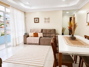 A modern living room with a sofa, a dining table, and a view through a glass door. - Aconchegante Apto Prainha Arraial- RJ (Arraial do Cabo)