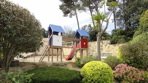 Ein Spielplatz mit Rutsche und Klettergerüst, umgeben von gepflegten Büschen und Bäumen.