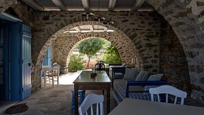 A stone-walled room with a table and chairs, a bench, and a view through an archway to a garden. - Belogna Ikons (Naxos)