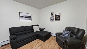 Een woonkamer met een hoekbank, een lederen fauteuil en een kleine bijzettafel. Er hangen drie ingelijste foto's aan de muur.