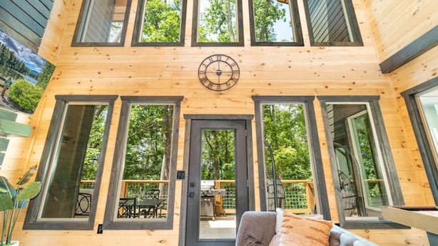 A wooden cabin with a large clock on the front door, multiple windows, and a cozy seating area with a table and chairs.