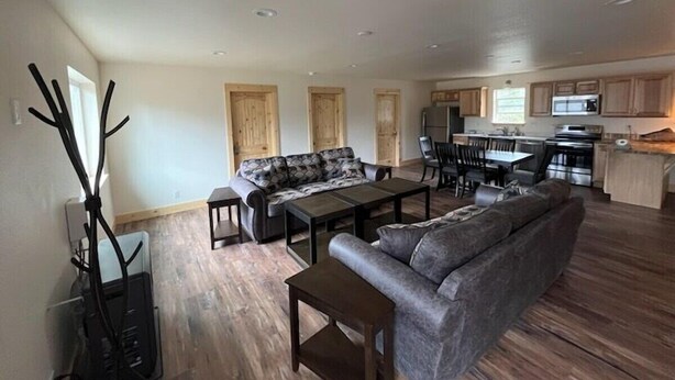 A spacious living area with a sofa, coffee table, and dining table. A kitchen with cabinets and appliances is visible in the background. - Alaska Summers Lodge Loft-Near Mouth of Kasilof River (Cohoe)
