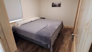 A small, narrow room with a single bed, a window with blinds, a picture on the wall, and a door. - Alaska Summers Lodge Loft-Near Mouth of Kasilof River (Cohoe)