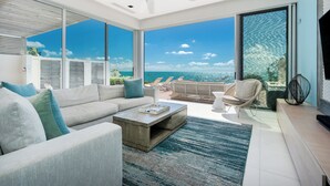A modern living room with a large window overlooking the ocean, a ceiling fan, and a seating area with a coffee table. - Beachfront Villa w/ Private Pool & Concierge – Villa Helios (Long Bay Beach)