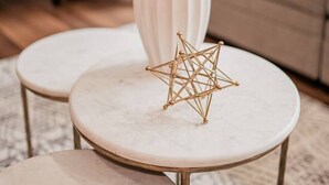 A living room with a blue chair, a beige sofa, and two round tables with a white vase and a gold geometric sculpture on one of them.