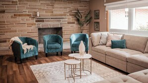 A living room with a stone wall, a fireplace, two teal armchairs, a beige sofa, and a small round table with a vase.