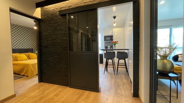 Un interior moderno con una puerta corredera que da a una zona de cocina, una cama con ropa de cama amarilla y un comedor con dos sillas.