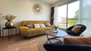 Una sala de estar moderna con un sofá, una mesita redonda y una silla. Hay un espejo decorativo en la pared y un florero con flores sobre una mesita auxiliar.