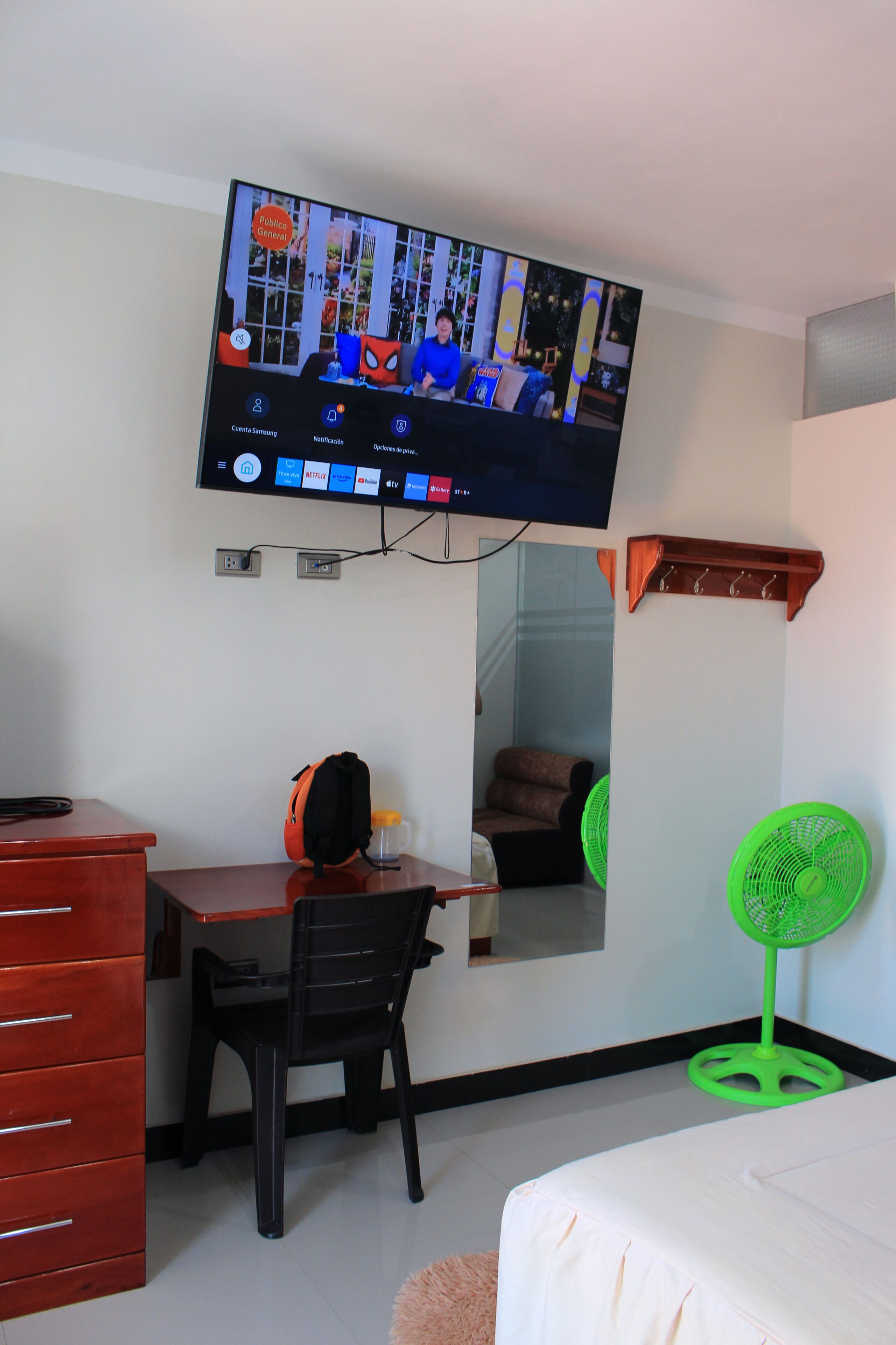 Une chambre d’hôtel équipée d’un téléviseur fixé au mur, diffusant une chaîne d’information, d’un bureau avec une chaise, d’un lit, d’une petite table avec une bouteille et d’un ventilateur vert.