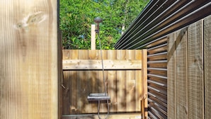 Ducha exterior de madera con una manguera, rodeada por una valla de madera y un portón metálico.
