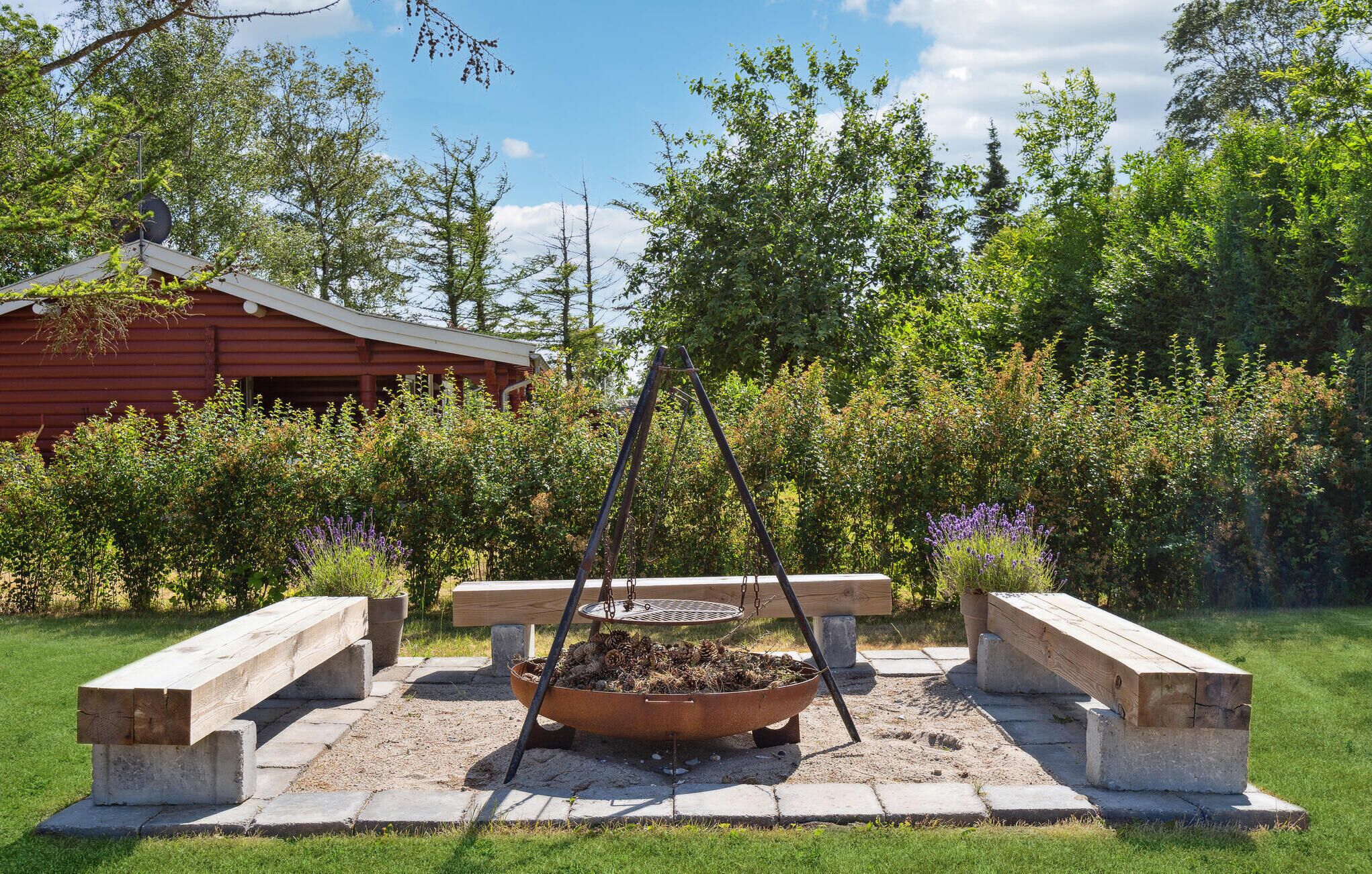A rustic outdoor seating area with a fire pit, surrounded by lush greenery and a wooden structure in the background.