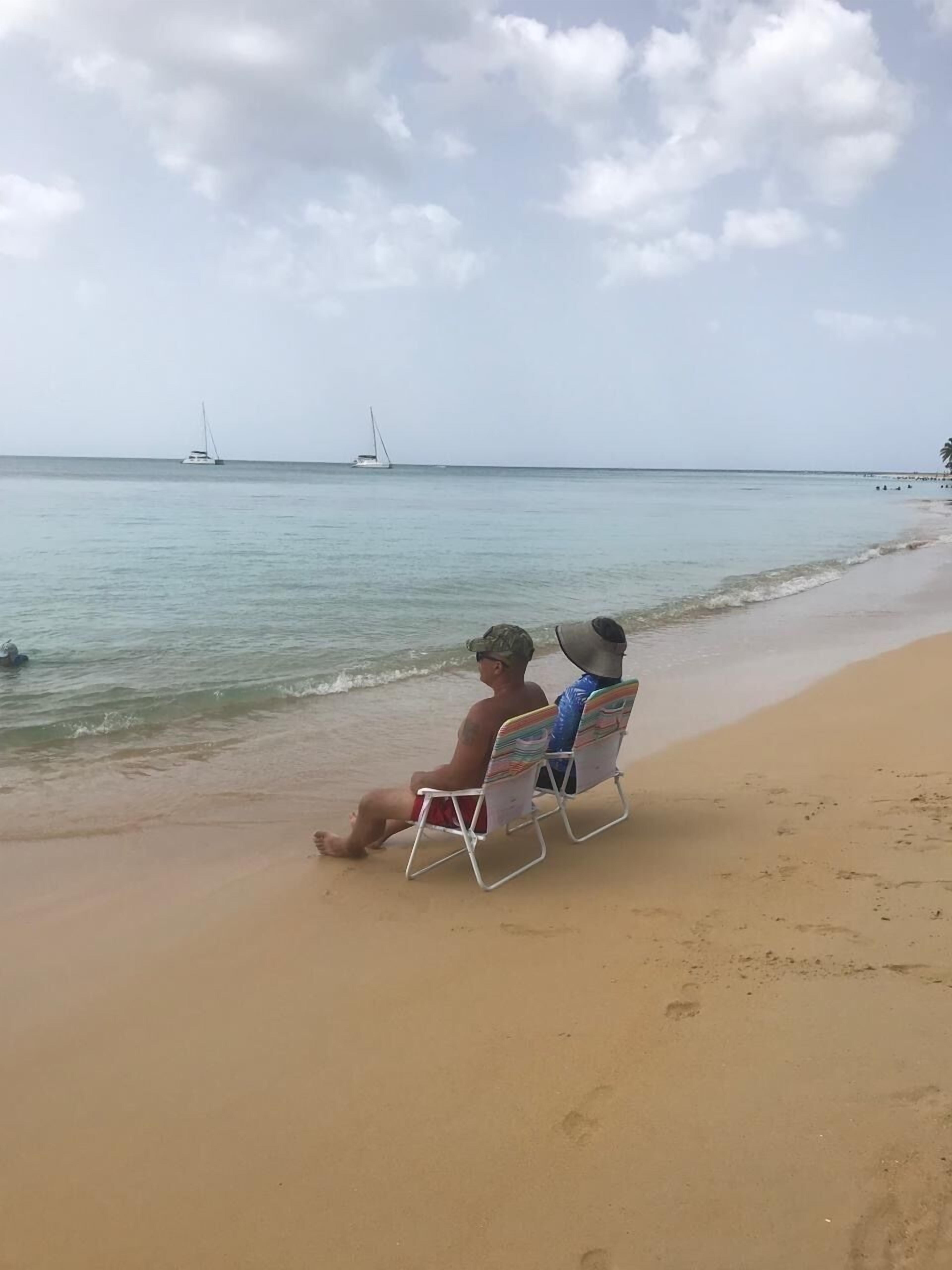 Zwei Personen sitzen auf Strandliegen und blicken aufs Meer, im Hintergrund ein Segelboot.