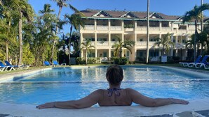 Eine Person entspannt am Pool mit Blick auf ein mehrstöckiges Gebäude mit Balkonen und Palmen.