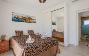 Une chambre à coucher comprenant un lit, une table de chevet, une commode, un grand miroir et un tableau représentant un paysage.
