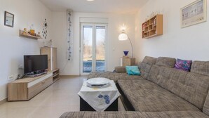 Een woonkamer met een bank, televisie en een tafel met een bord. Er is een deur die naar buiten leidt en een plank met decoratieve objecten.