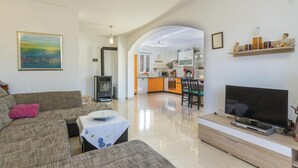 Een woonkamer met een bank, een televisie en een open haard. In de achtergrond is een keuken zichtbaar.