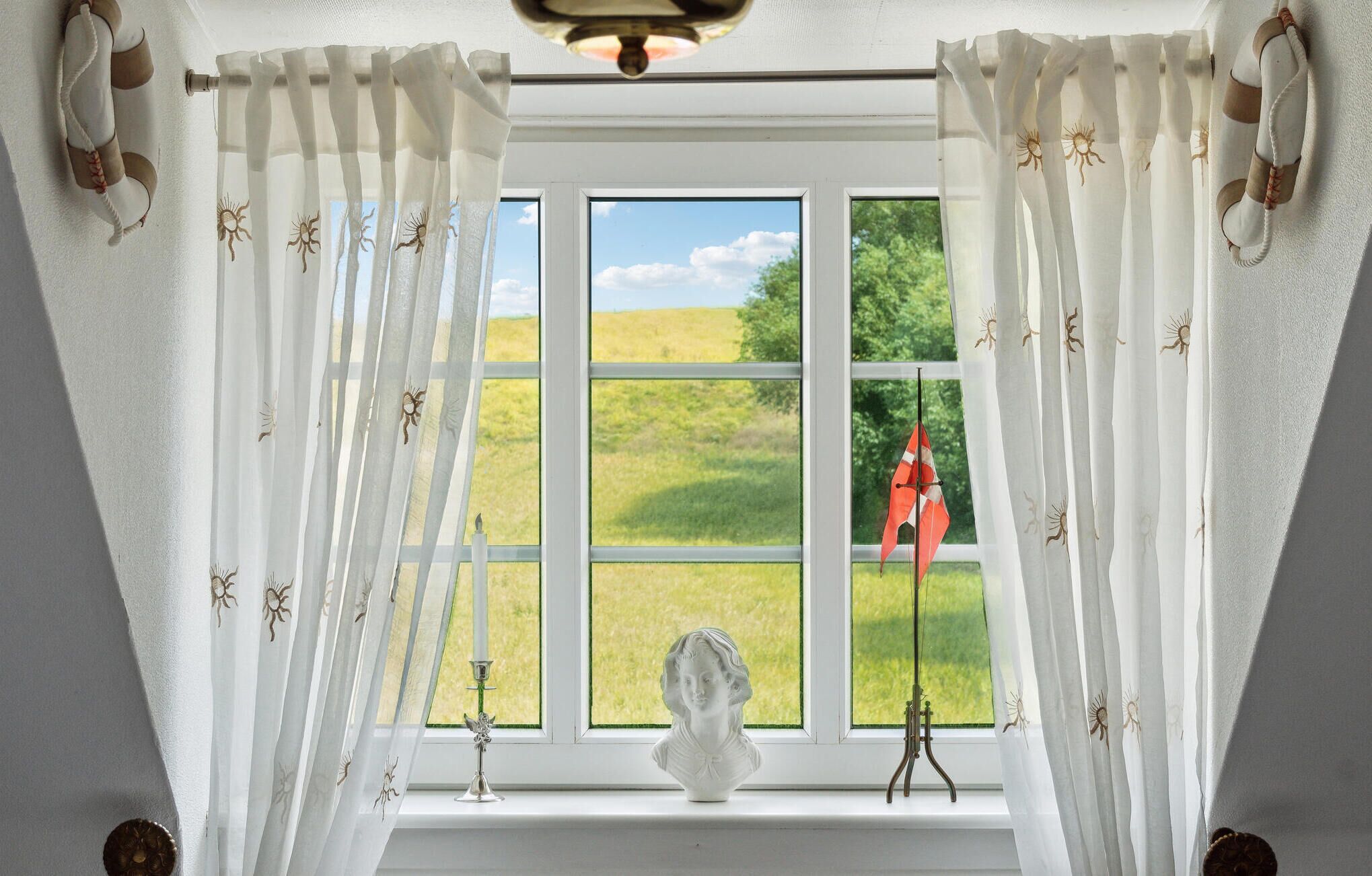 A room with a large window, white curtains with a pattern, a bust sculpture, and a flag outside.
