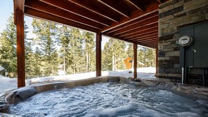 Un espace avec un bain à remous, doté d’un mur en pierre et de poutres en bois, entouré de neige et d’arbres.