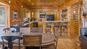 A rustic cabin interior with a wooden dining table, bar stools, a kitchen with wooden cabinets, a refrigerator, and a large wooden tree trunk. - Gorgeous Unique, Rustic Cabin on Arapaho! 2 Kings, Loft Area with a Full, and a Bunk Room! (Duck Creek Village)