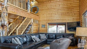 A log cabin interior with a wooden staircase, a sectional sofa, a grey armchair, and a small table with a lamp. - Gorgeous Unique, Rustic Cabin on Arapaho! 2 Kings, Loft Area with a Full, and a Bunk Room! (Duck Creek Village)