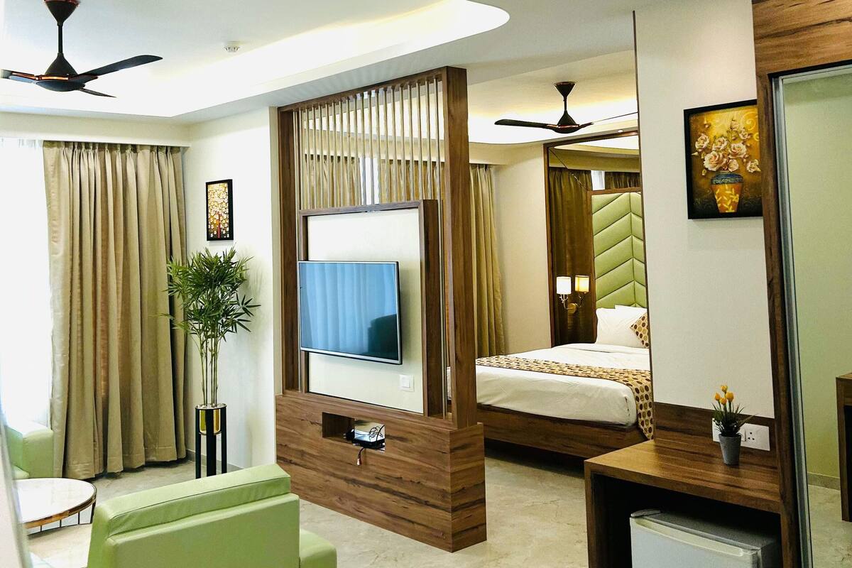 Una habitación de hotel moderna con televisor de pantalla plana, cama con colcha estampada, sofá verde, refrigerador pequeño y ventilador de techo.