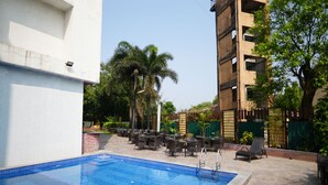Una piscina con sillones y mesas, rodeada de follaje y un edificio de varios pisos.