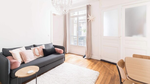 Un salon moderne avec un canapé, une petite table ronde, un tapis blanc, une table à manger en bois et un lustre.