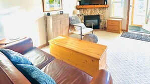 A living room with a brown leather sofa, a wooden coffee table, a stone fireplace, a television, and a ceiling fan. - Perfect Creekside Studio that Opens Right onto the Skiway!  (Silver Star Mountain)