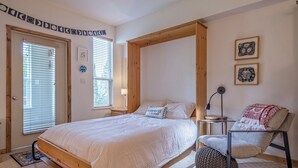 A bedroom with a wooden bed frame, a bedside table, a chair, and two framed pictures on the wall. - Perfect Creekside Studio that Opens Right onto the Skiway!  (Silver Star Mountain)