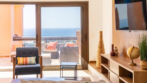Ein Wohnzimmer mit einer Couch, einem kleinen Tisch, einem Fernseher und Blick auf den Ozean durch eine Schiebetür.