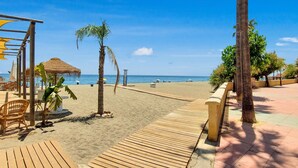 Ein hölzerner Steg führt zu einem Strand mit Reetsonnenstühlen und Palmen.