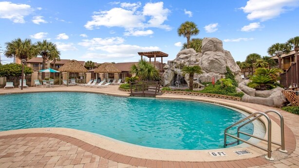 A swimming pool with a rock formation and palm trees, surrounded by lounge chairs and a gazebo.
