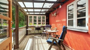Un porche cubierto con piso de madera, una parrilla, una mesa con sillas y una pared roja con una ventana.