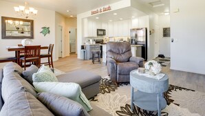 A spacious living room with a grey sofa, a brown leather chair, and a small round table with flowers. A kitchen is visible in the background with white cabinets and modern appliances. - Mesquite Vacation Rental Condo w/ Resort Amenities (Mesquite)