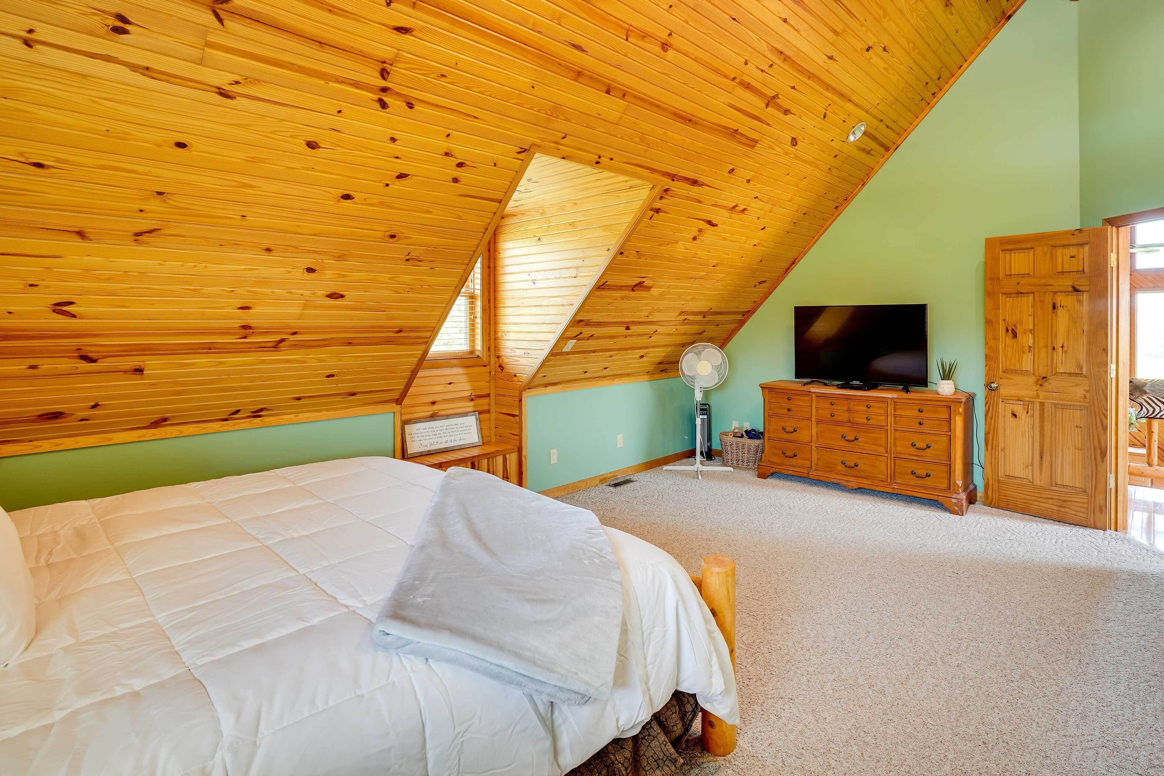 A bedroom with a sloped wooden ceiling, a bed, a dresser, a television, and a fan.