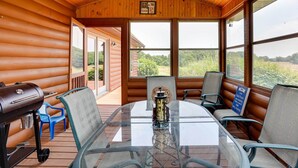 Interior de uma cabana de madeira com mesa de vidro, cadeiras, churrasqueira e vista para o exterior.